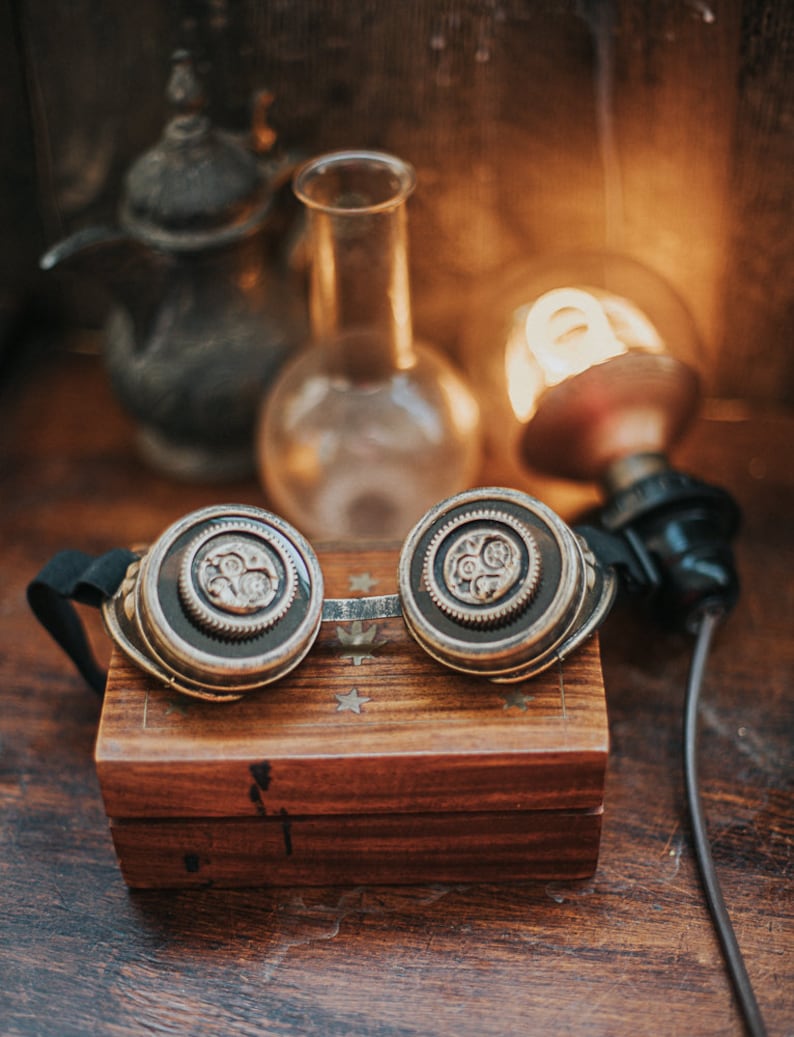 Puede incluir: Un par de gafas steampunk con detalles en lat&oacute;n y plata, descansando sobre una caja de madera con un patr&oacute;n de estrellas. Las gafas tienen una correa de cuero negro y est&aacute;n enfocadas, mientras que el fondo est&aacute; desenfocado.