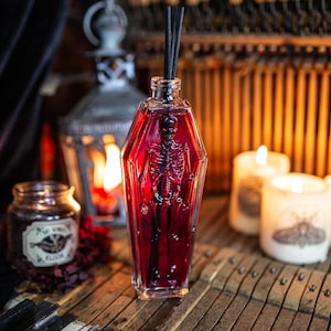 May include: A glass bottle shaped like a coffin filled with a red liquid and a skeleton inside. The bottle has black reeds sticking out of the top. The bottle is sitting on a wooden surface with a piano keyboard in the foreground.
