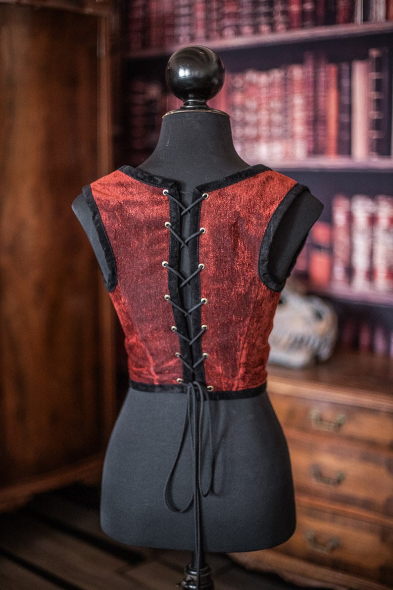 Op de afbeelding: Een rood fluwelen korset met zwarte vetersluiting aan de achterkant. Het korset is mouwloos en heeft een strak lijfje.