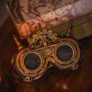 May include: A pair of brass-colored goggles featuring two circular dark lenses, a central gear mechanism, and small protruding antenna-like components on each side. The frame has a metallic finish with intricate detailing around the eye cups.