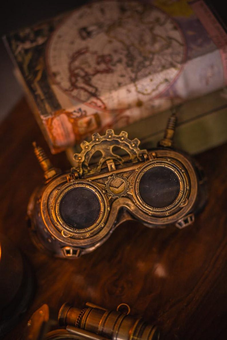 May include: A pair of brass-colored goggles featuring two circular dark lenses, a central gear mechanism, and small protruding antenna-like components on each side. The frame has a metallic finish with intricate detailing around the eye cups.