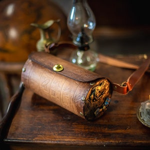 May include: A brown leather cylindrical bag with an intricate design and gold accents. The bag has a strap and is sitting on a wooden table.