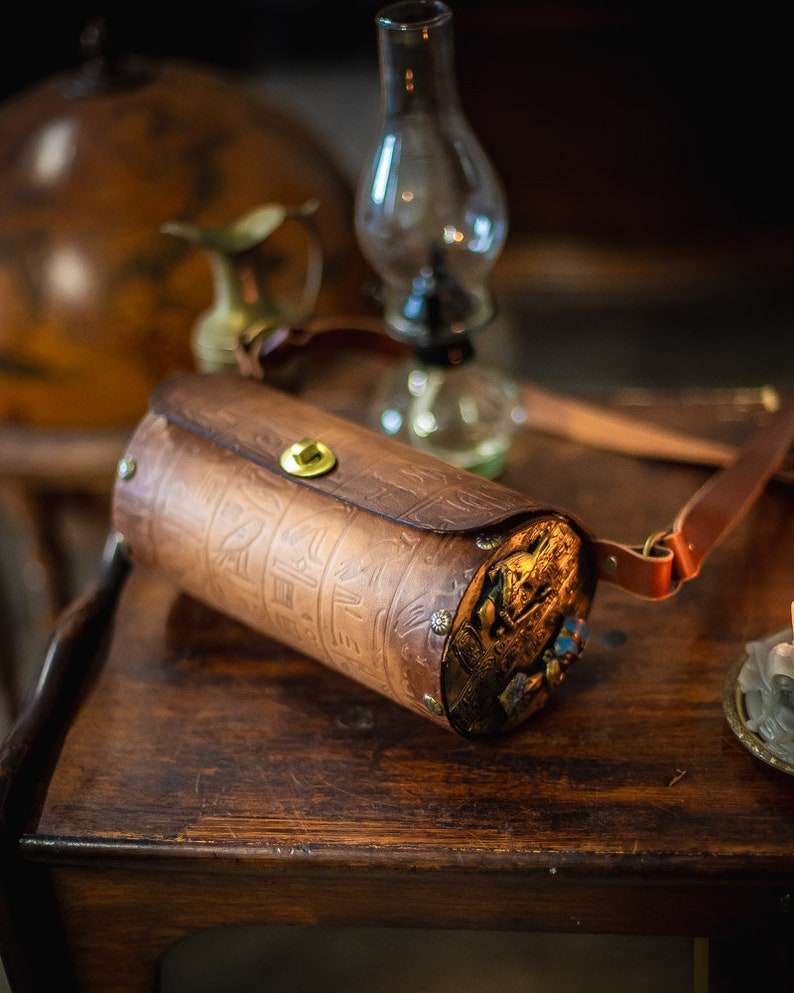 May include: A brown leather cylindrical bag with an intricate design and gold accents. The bag has a strap and is sitting on a wooden table.
