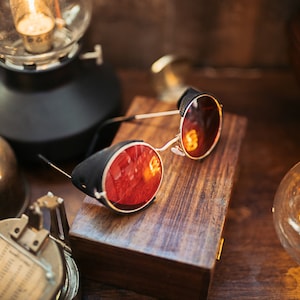 May include: Round red-tinted steampunk goggles with black leather accents on a wooden box. The goggles are on a wooden surface with a vintage light bulb and other antique objects.