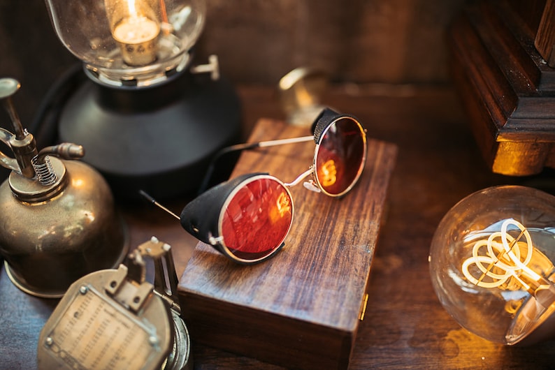 May include: Round red-tinted steampunk goggles with black leather accents on a wooden box. The goggles are on a wooden surface with a vintage light bulb and other antique objects.