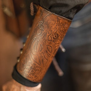 May include: Close-up of a brown leather arm guard with intricate embossed designs. The arm guard features a series of floral and abstract patterns. The leather has a distressed finish, giving it a vintage look. The arm guard is worn over a black sleeve.