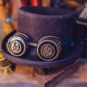 Puede incluir: Un sombrero de copa negro con un par de gafas steampunk. Las gafas tienen lentes redondas de color lat&oacute;n con intrincados dise&ntilde;os de engranajes.
