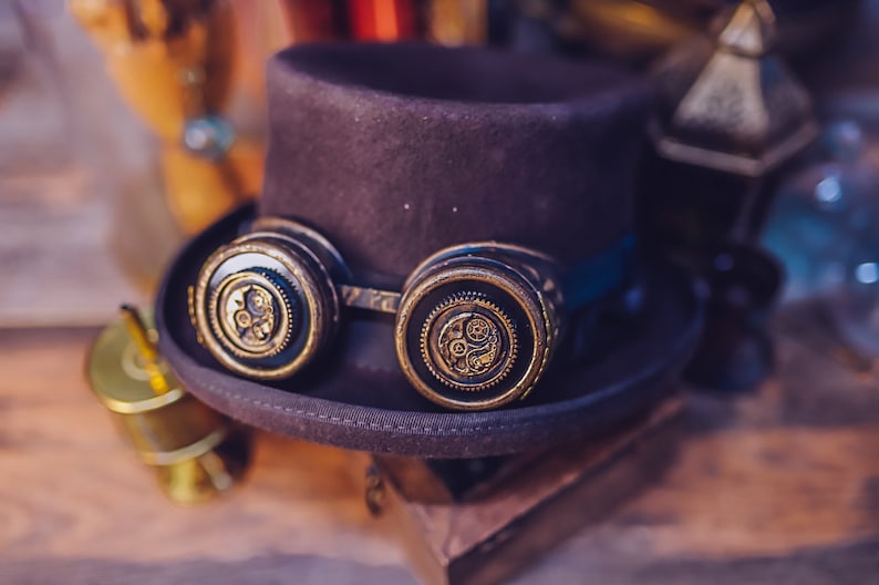 Puede incluir: Un sombrero de copa negro con un par de gafas steampunk. Las gafas tienen lentes redondas de color lat&oacute;n con intrincados dise&ntilde;os de engranajes.