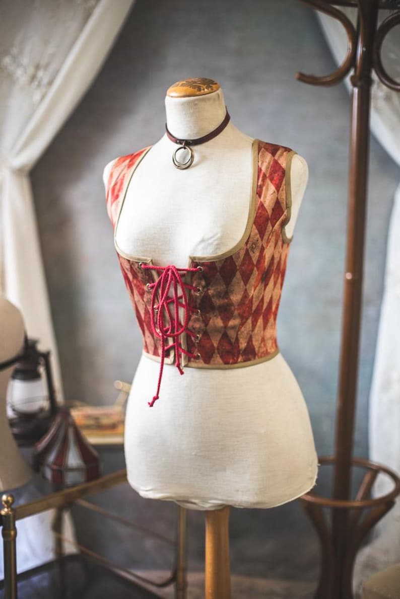 May include: A vintage-style corset displayed on a mannequin. The corset features a red and tan diamond pattern, a red lace-up front, and a brown trim. A burgundy choker necklace with a silver pendant is also visible.