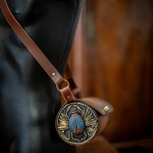 May include: A brown leather roll with a decorative metal clasp featuring a blue and red scarab beetle with gold wings. The roll is worn over a shoulder strap.