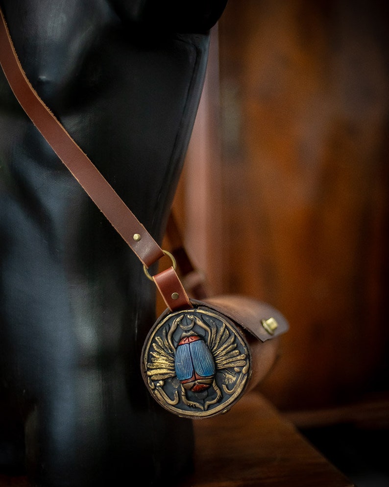 May include: A brown leather roll with a decorative metal clasp featuring a blue and red scarab beetle with gold wings. The roll is worn over a shoulder strap.