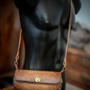 May include: A brown leather crossbody bag with a brass clasp and decorative studs. The bag has an embossed pattern of hieroglyphics.