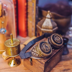 Puede incluir: Un par de gafas de estilo steampunk con marcos de lat&oacute;n y correas de cuero negro. Las gafas est&aacute;n sobre una caja de madera con un soporte decorativo de metal dorado y una c&uacute;pula de vidrio que contiene una mano de madera.