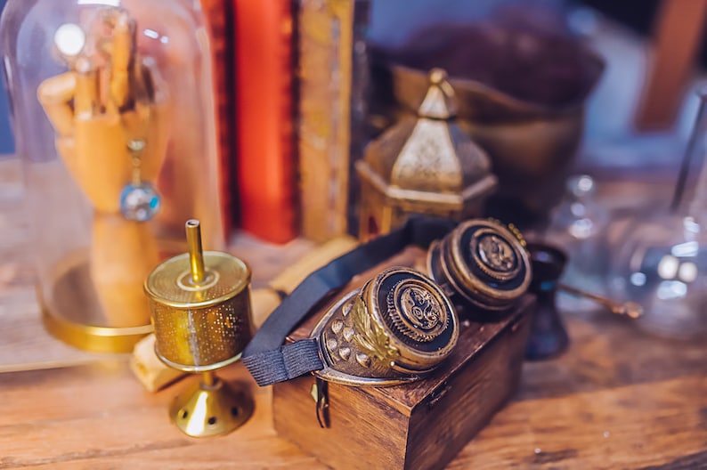 Puede incluir: Un par de gafas de estilo steampunk con marcos de lat&oacute;n y correas de cuero negro. Las gafas est&aacute;n sobre una caja de madera con un soporte decorativo de metal dorado y una c&uacute;pula de vidrio que contiene una mano de madera.