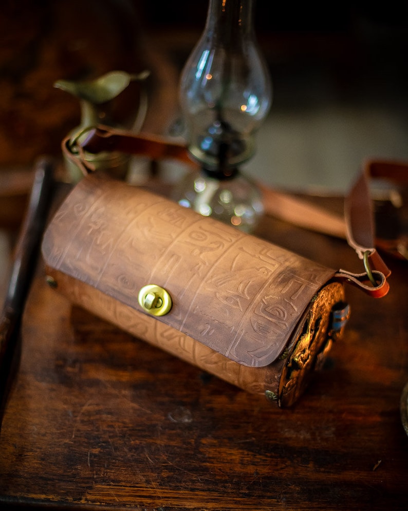 May include: A brown leather cylindrical bag with an embossed design and a brass clasp. The bag has a long brown leather strap.