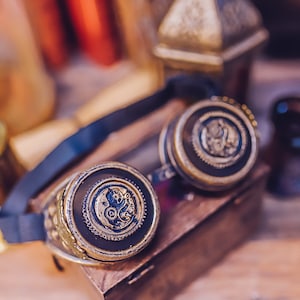 Puede incluir: Un par de gafas steampunk con una correa de cuero negro y detalles met&aacute;licos dorados. Las gafas tienen lentes redondas con intrincados dise&ntilde;os de relojer&iacute;a.