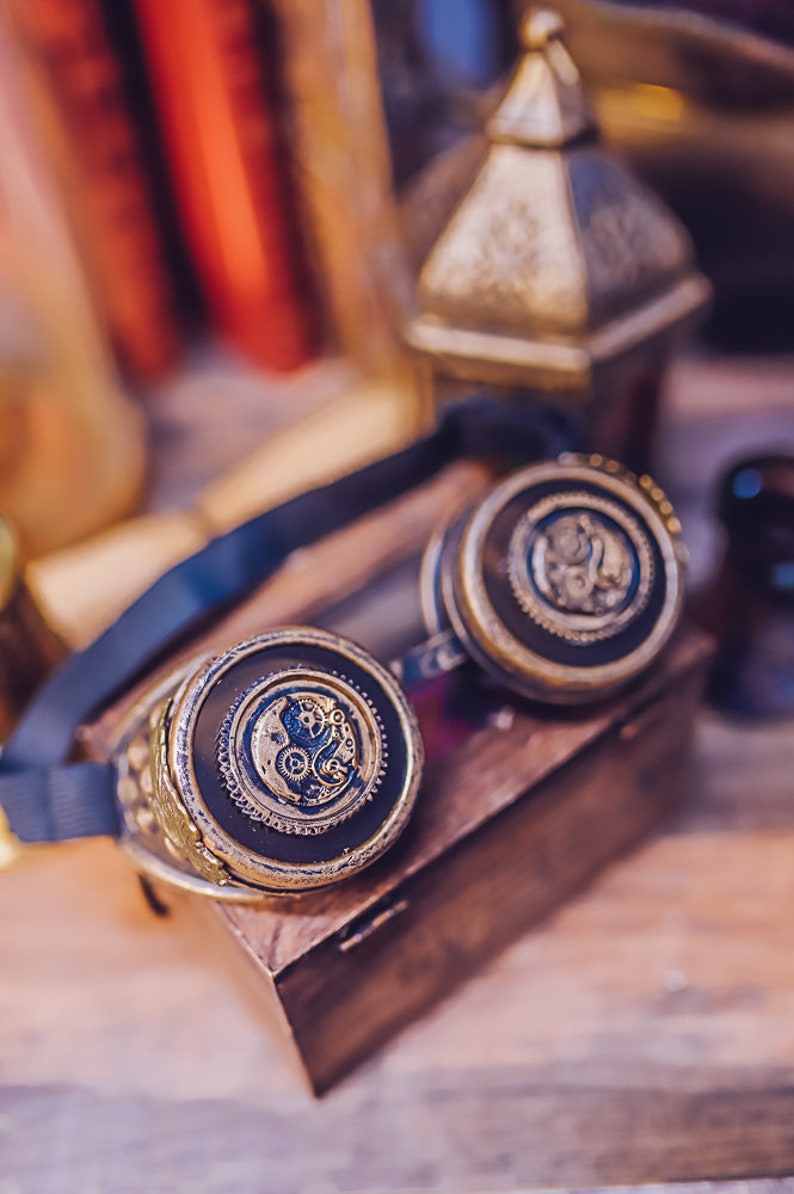 Puede incluir: Un par de gafas steampunk con una correa de cuero negro y detalles met&aacute;licos dorados. Las gafas tienen lentes redondas con intrincados dise&ntilde;os de relojer&iacute;a.