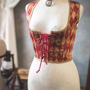 May include: A corset displayed on a mannequin. The corset features a red and tan diamond pattern, a red lace-up front, and a brown trim. A matching choker with a bronze ring is around the mannequin's neck. The corset is a vintage style.