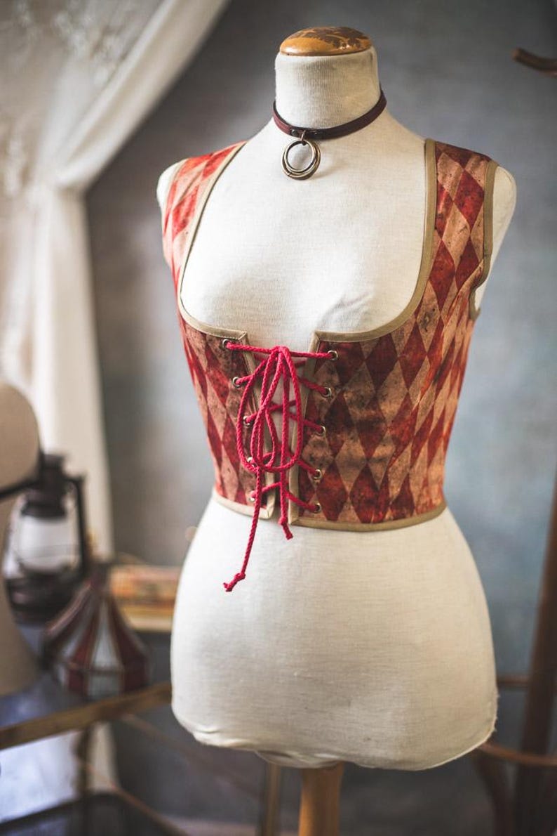 May include: A corset displayed on a mannequin. The corset features a red and tan diamond pattern, a red lace-up front, and a brown trim. A matching choker with a bronze ring is around the mannequin's neck. The corset is a vintage style.
