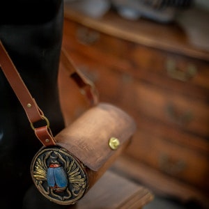 May include: A brown leather cylindrical bag with a gold-colored metal clasp and a decorative circular panel featuring a blue and gold scarab beetle design.