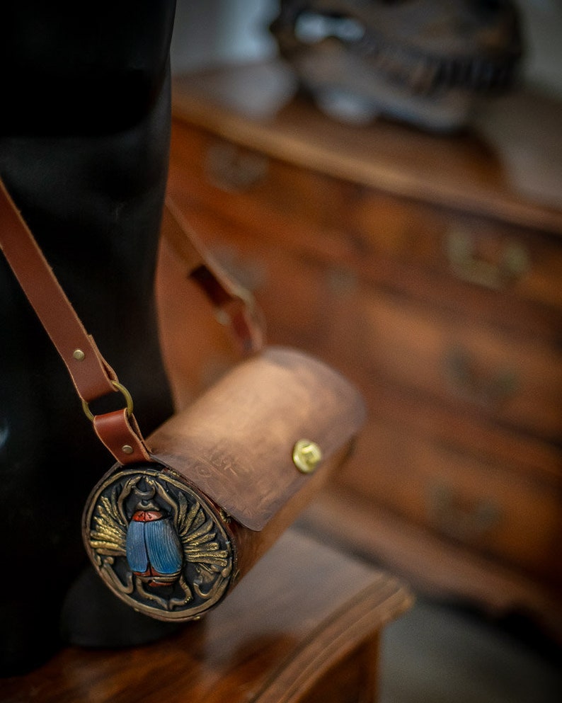 May include: A brown leather cylindrical bag with a gold-colored metal clasp and a decorative circular panel featuring a blue and gold scarab beetle design.