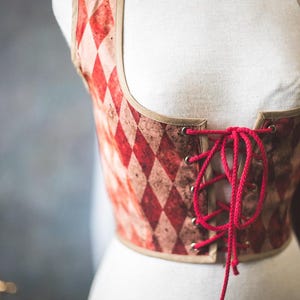 May include: A red and white diamond-patterned corset with a red lace-up front. The corset has a tan trim and is displayed on a mannequin. A brown choker with two gold rings is around the mannequin's neck.