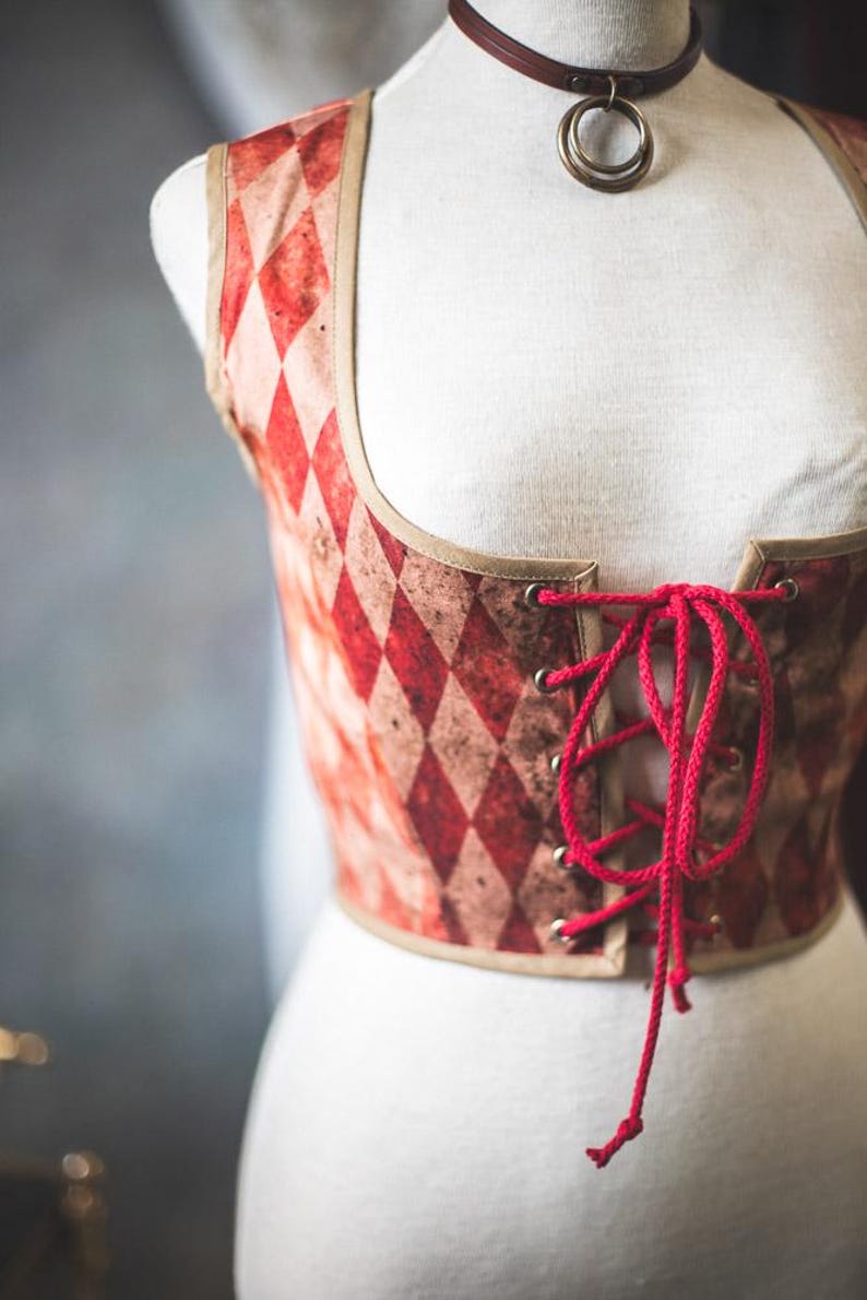 May include: A red and white diamond-patterned corset with a red lace-up front. The corset has a tan trim and is displayed on a mannequin. A brown choker with two gold rings is around the mannequin's neck.