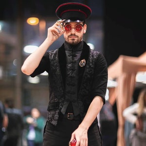 May include: A man wearing a black and white patterned vest, a black shirt, and black pants. He is wearing a black and red military-style hat and red sunglasses. He is holding a red and gold object in his right hand.