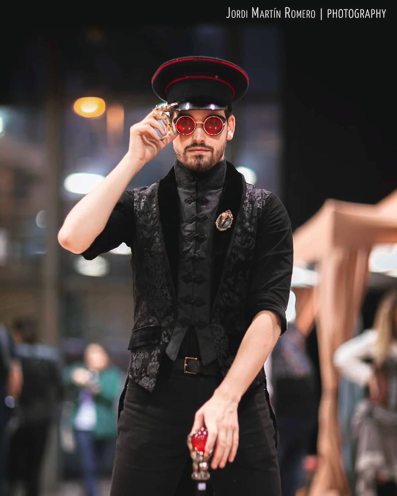 May include: A man wearing a black and white patterned vest, a black shirt, and black pants. He is wearing a black and red military-style hat and red sunglasses. He is holding a red and gold object in his right hand.