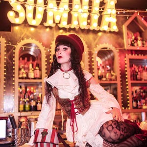 May include: A person in a vintage-inspired outfit poses in front of a bar with the word "SOMNIA" in illuminated letters. The outfit includes a red hat, a white dress, and a brown corset. A small, striped bag sits on the bar.