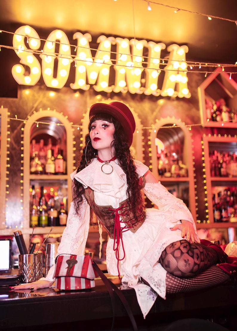 May include: A person in a vintage-inspired outfit poses in front of a bar with the word "SOMNIA" in illuminated letters. The outfit includes a red hat, a white dress, and a brown corset. A small, striped bag sits on the bar.