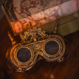 May include: A pair of brass-colored goggles featuring two circular dark lenses, a central gear mechanism, and two small vertical antennas on the sides. The frame is detailed with metallic ridges and circular markings around the lens rims.