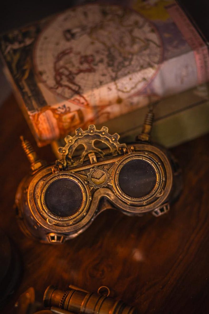 May include: A pair of brass-colored goggles featuring two circular dark lenses, a central gear mechanism, and two small vertical antennas on the sides. The frame is detailed with metallic ridges and circular markings around the lens rims.