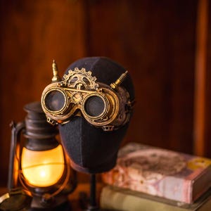 May include: Brass-colored goggles featuring circular lenses, a central gear-shaped ornament, and two small vertical spikes on the top frame. The goggles are mounted on a dark, featureless mannequin head stand, showcasing intricate metallic details.