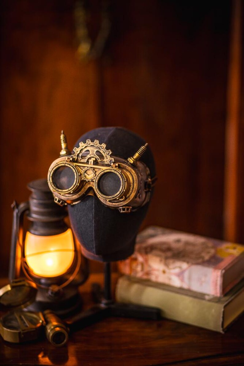 May include: Brass-colored goggles featuring circular lenses, a central gear-shaped ornament, and two small vertical spikes on the top frame. The goggles are mounted on a dark, featureless mannequin head stand, showcasing intricate metallic details.