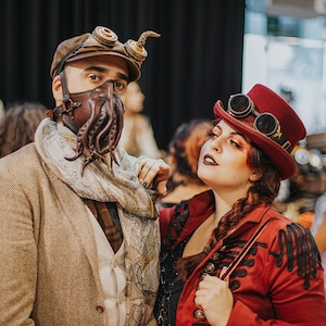 May include: Two people dressed in steampunk attire. The person on the left is wearing a brown jacket, a scarf, and a leather mask with tentacles. The person on the right is wearing a red jacket, a red hat with goggles, and dark makeup.