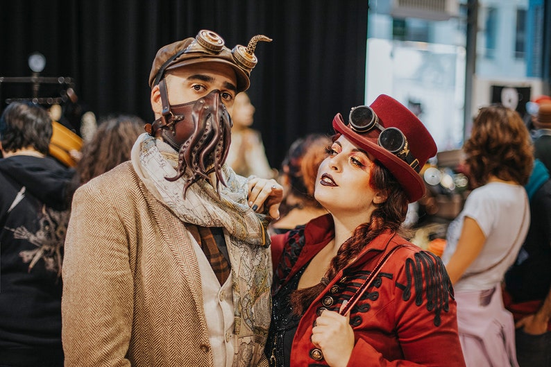 May include: Two people dressed in steampunk attire. The person on the left is wearing a brown jacket, a scarf, and a leather mask with tentacles. The person on the right is wearing a red jacket, a red hat with goggles, and dark makeup.