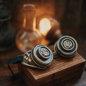 Puede incluir: Un par de gafas steampunk con detalles en lat&oacute;n y una correa negra. Las gafas est&aacute;n sobre una caja de madera con un dise&ntilde;o de estrella.