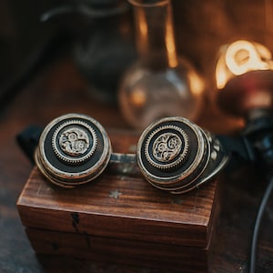 Puede incluir: Un par de gafas steampunk con marcos de color lat&oacute;n y dise&ntilde;os intrincados de engranajes en las lentes. Las gafas est&aacute;n apoyadas en una caja de madera.
