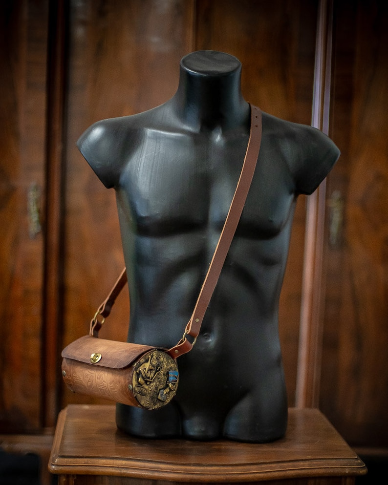May include: A brown leather cylindrical crossbody bag with a gold metal clasp and a decorative design. The bag is worn on a black mannequin.