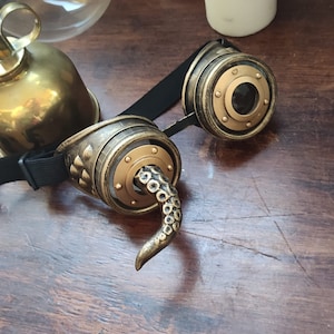 May include: A pair of steampunk-style goggles with a gold and black finish. The goggles have a round lens and a decorative tentacle detail on one side.