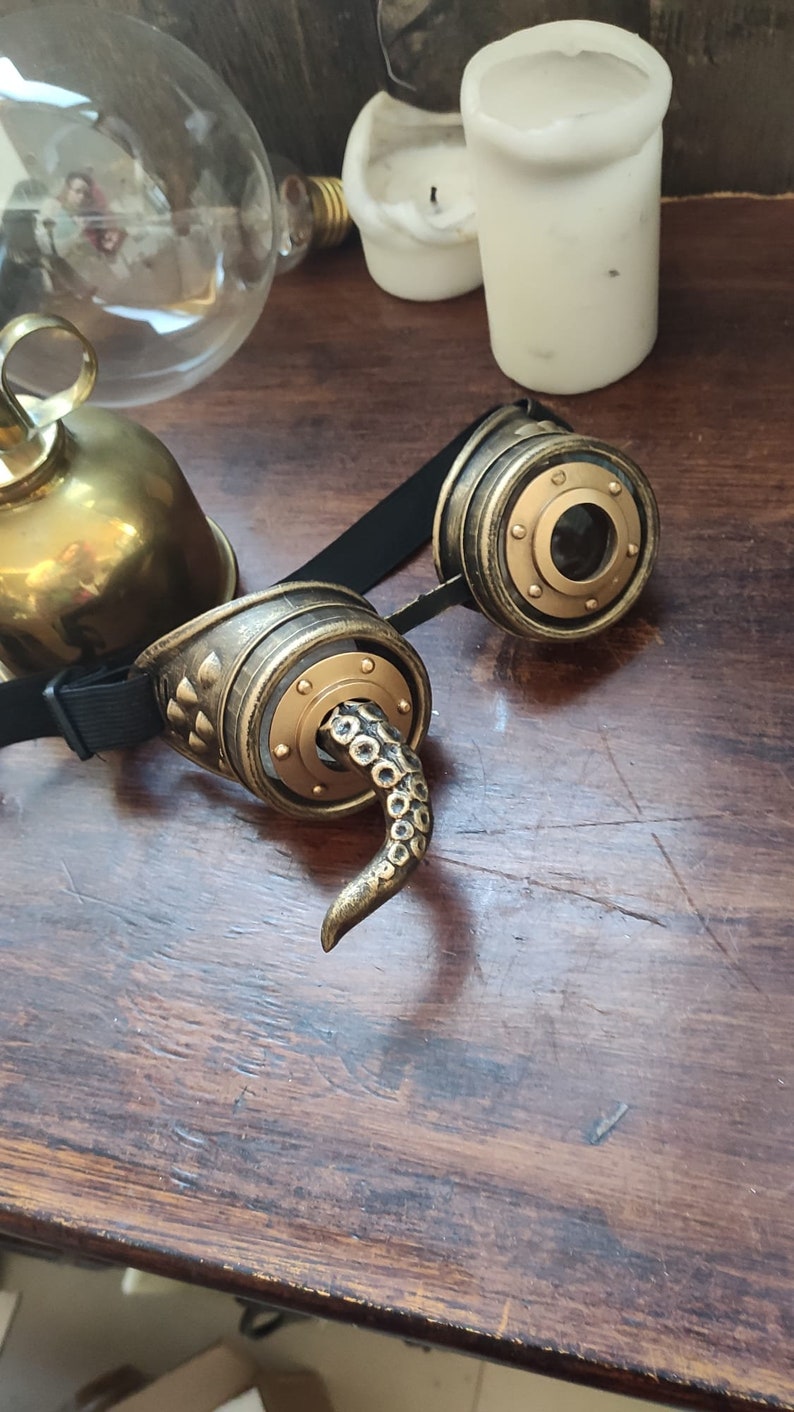 May include: A pair of steampunk-style goggles with a gold and black finish. The goggles have a round lens and a decorative tentacle detail on one side.