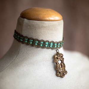 May include: A green and gold choker necklace with a keyhole pendant. The necklace is made of woven fabric and metal.