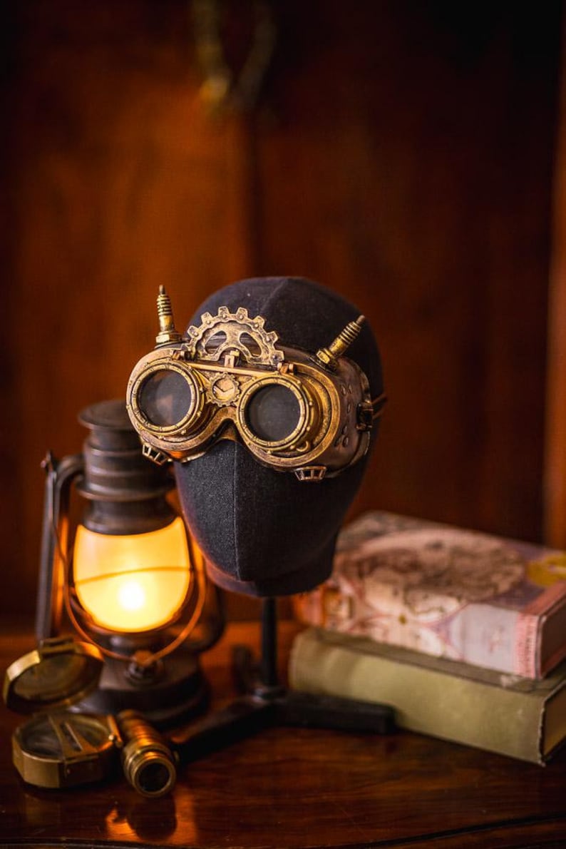 May include: Gold-toned brass goggles featuring a prominent gear mechanism on the bridge, two circular lenses, and small vertical spikes on the sides. The goggles are displayed on a dark fabric-covered head form, showcasing intricate metal detailing.