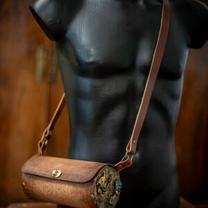 May include: A brown leather cylindrical crossbody bag with a brass buckle and decorative metal accents. The bag has a long brown leather strap.
