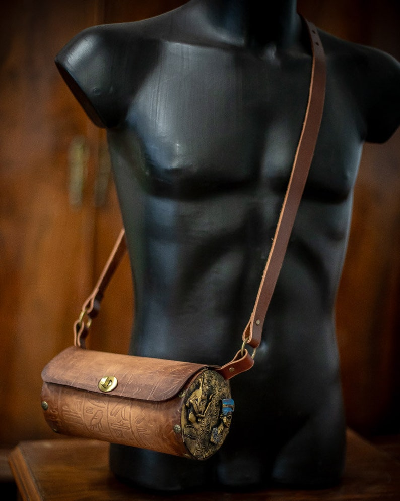 May include: A brown leather cylindrical crossbody bag with a brass buckle and decorative metal accents. The bag has a long brown leather strap.