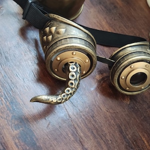 May include: A pair of steampunk goggles with a gold finish and an octopus tentacle detail. The goggles have a black adjustable strap.