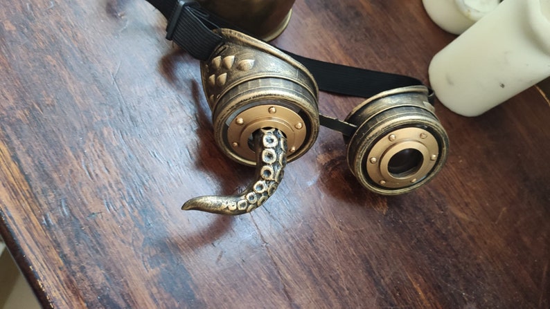May include: A pair of steampunk goggles with a gold finish and an octopus tentacle detail. The goggles have a black adjustable strap.