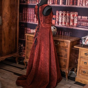 Op de afbeelding: Een rode fluwelen jurk met een zwarte veter aan de achterkant en een lange, zwierige rok. De jurk wordt getoond op een mannequin voor een boekenkast vol boeken.