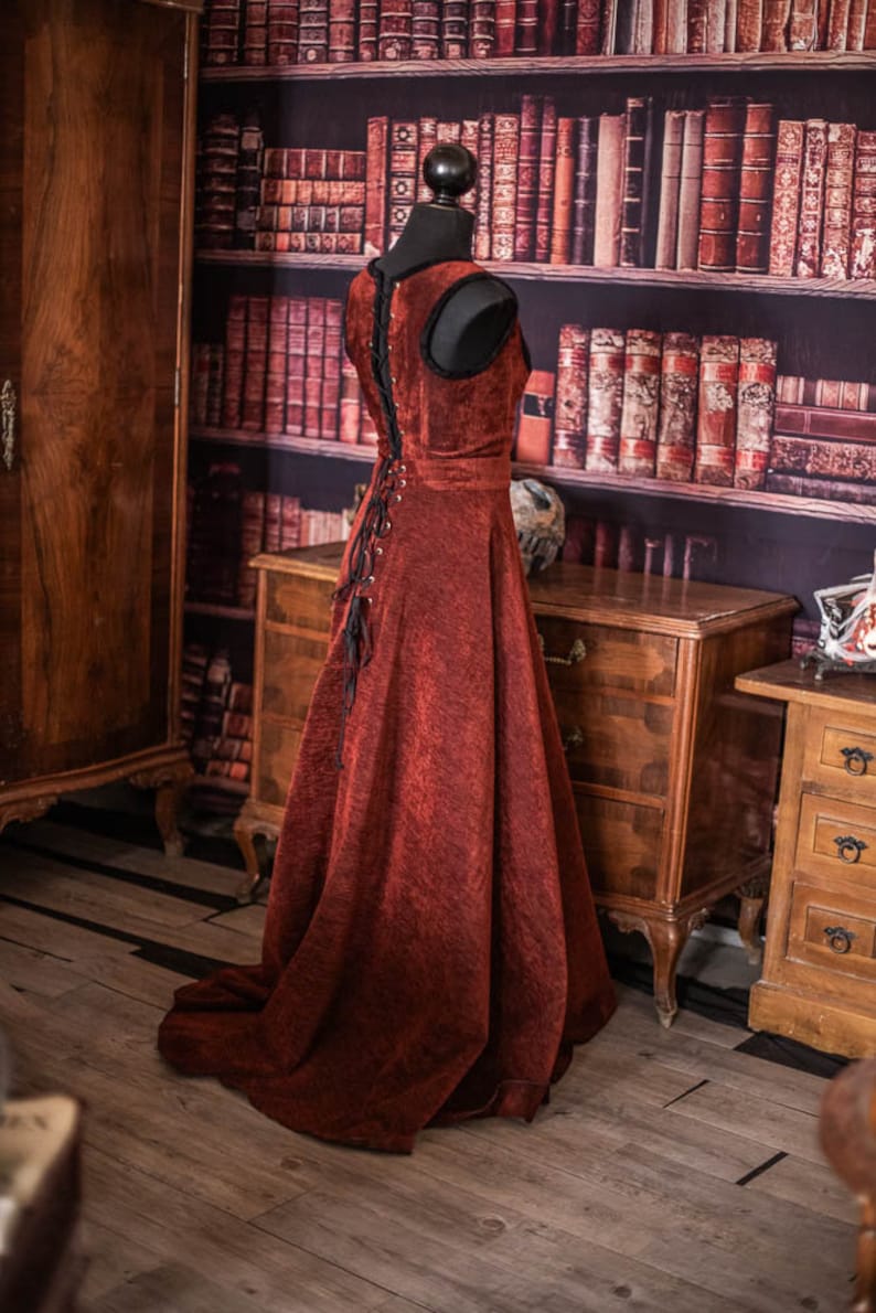 Op de afbeelding: Een rode fluwelen jurk met een zwarte veter aan de achterkant en een lange, zwierige rok. De jurk wordt getoond op een mannequin voor een boekenkast vol boeken.